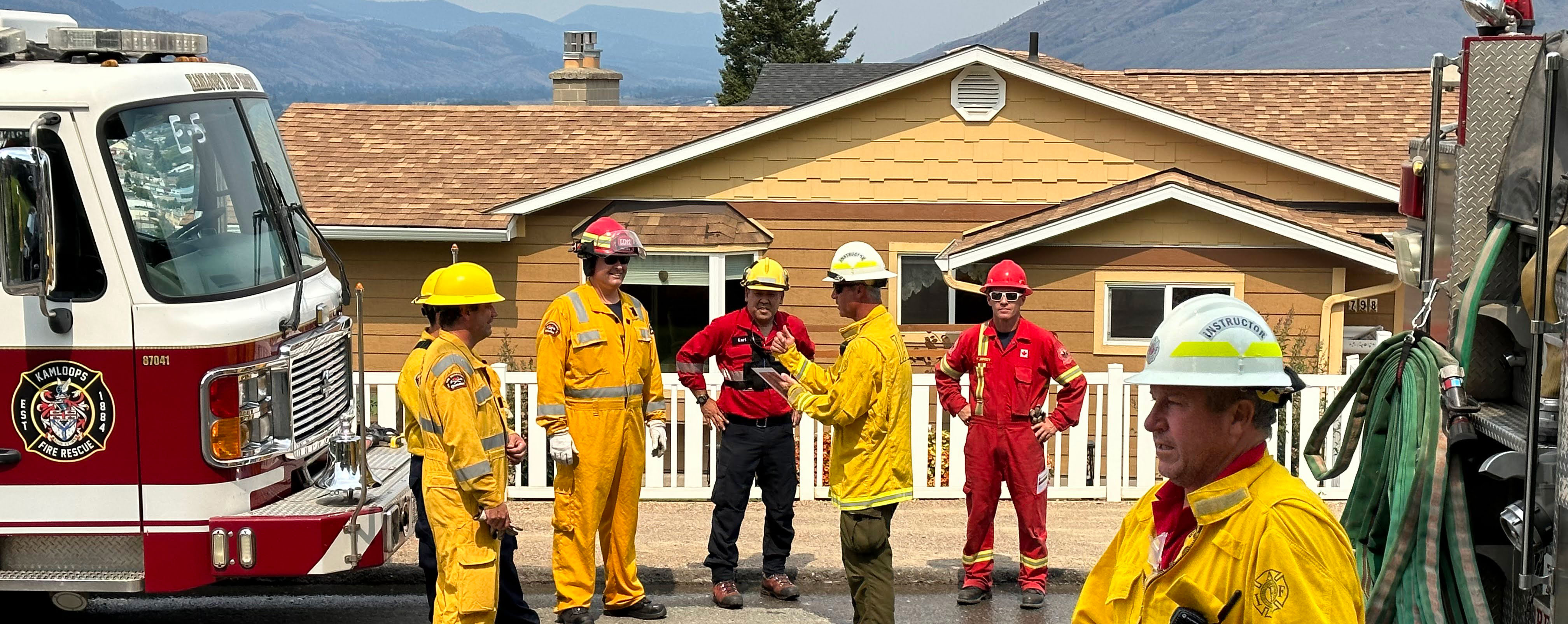 Six firefighters in front of a yellow house on the street listening to instructions