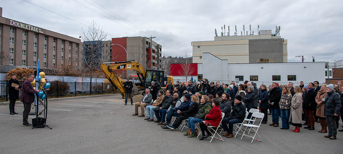 crowd standing and in chairs in an outdoor setting listening to a speech