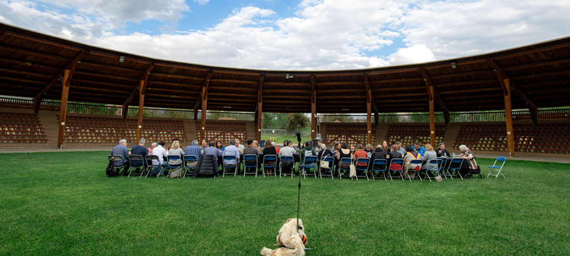 People seated at a long table in the centre of the open air powwow arbour located at Tḱemlúps te Secwépemc.