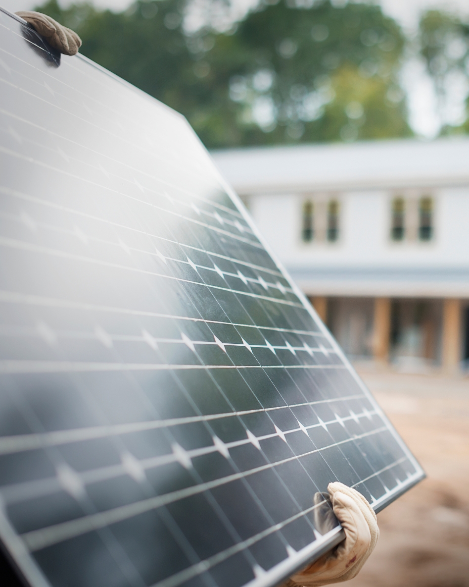 Two hands are holding up a solar panel in front of a house.