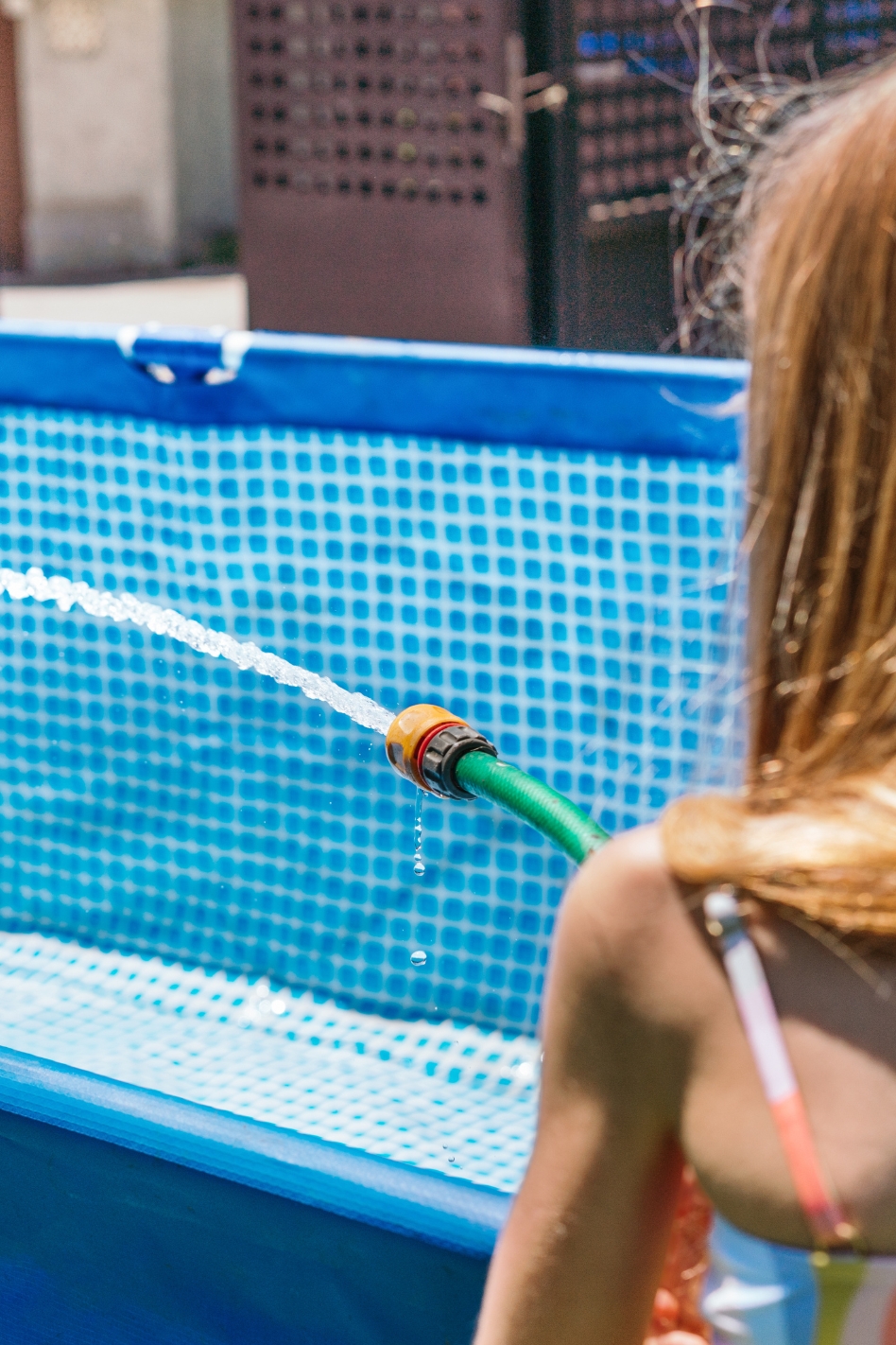 A young child points a hose with running water into an above-ground swimming pool.