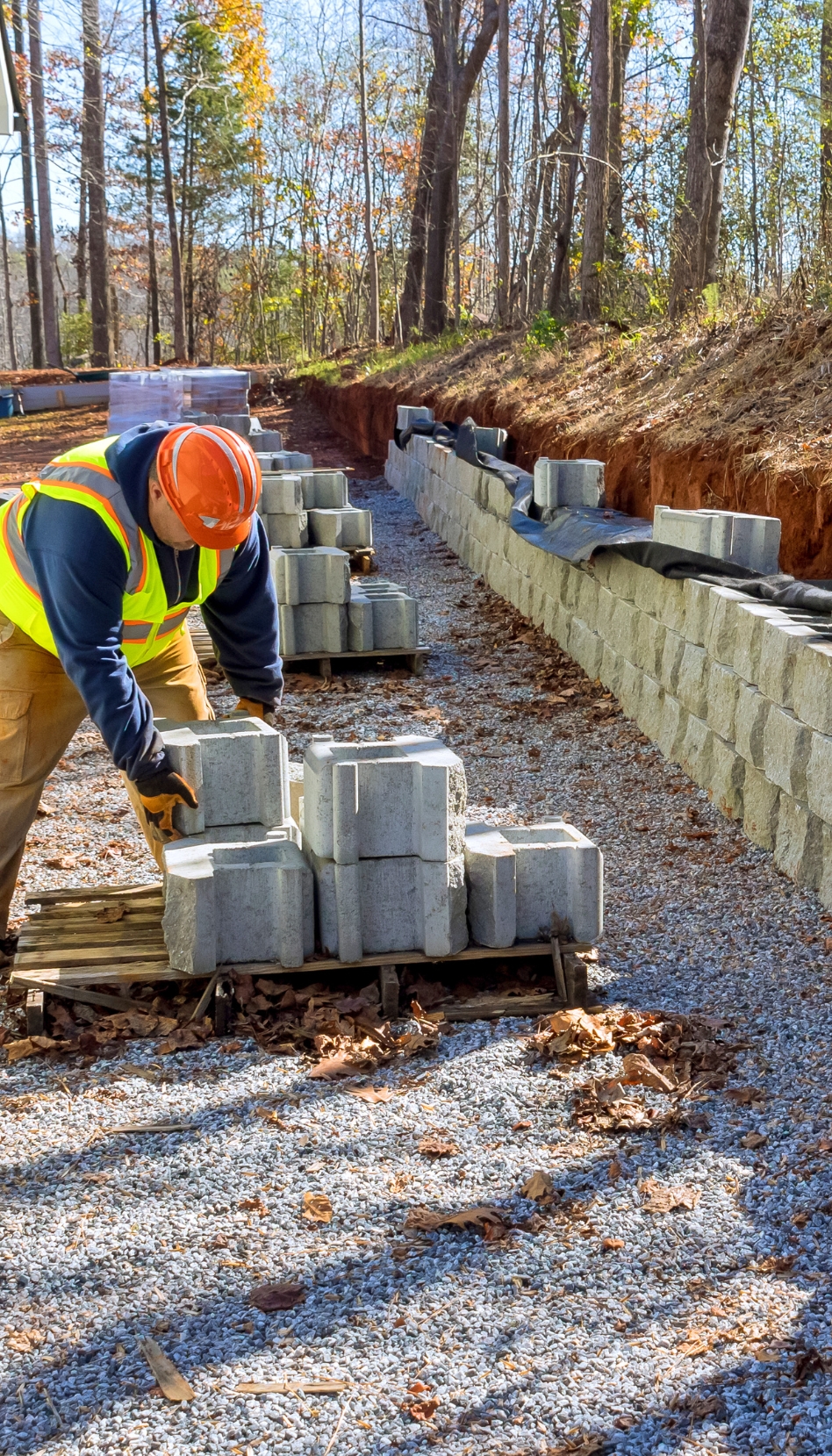 A contractor in a hard had and high visibility vest leans over a pallet of cinder blocks beside a retaining wall.