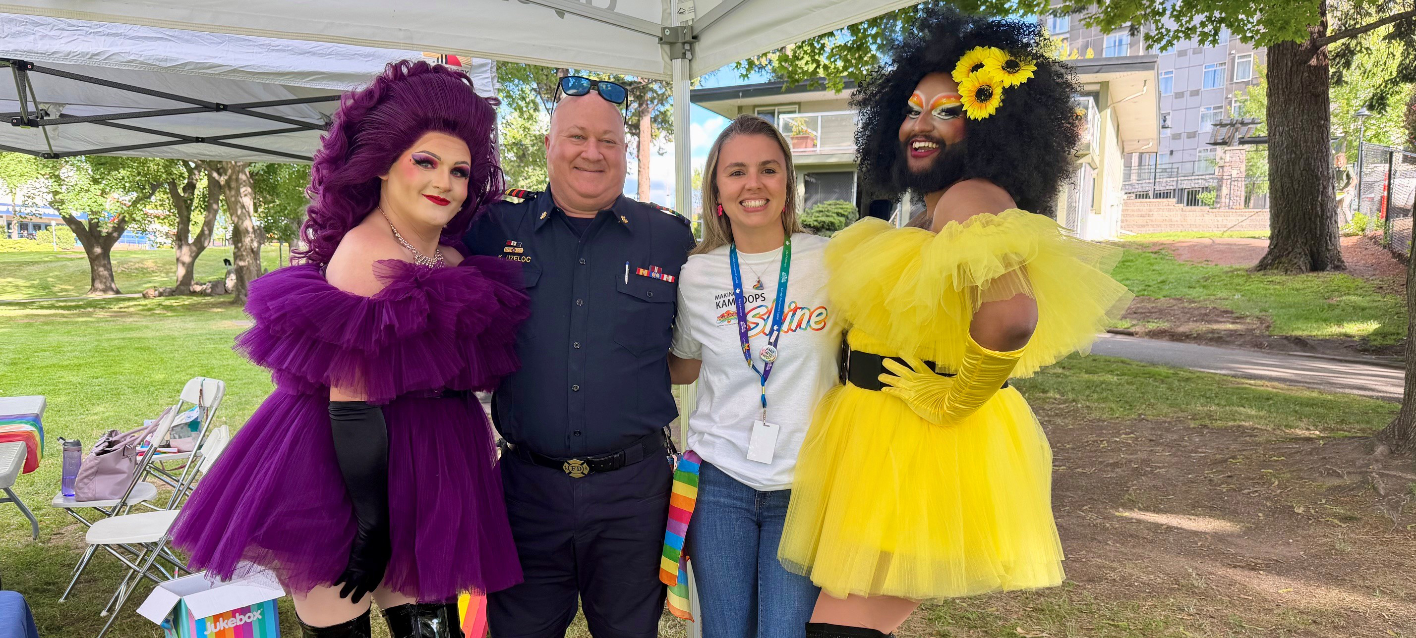 Two drag queens in matching bright fuchsia and yellow dresses pose and smile with Fire Chief and City Staffer at Riverside Park