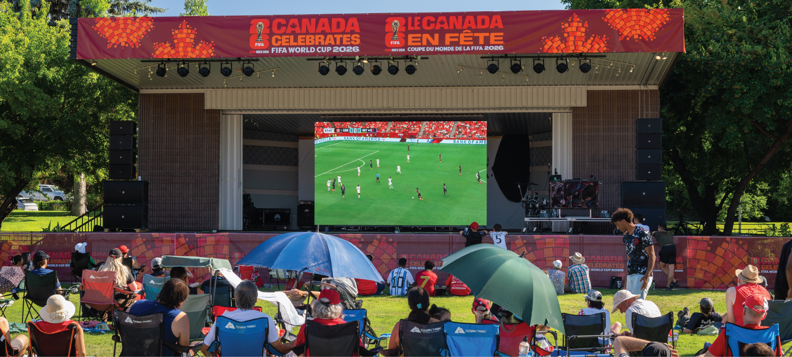 a big crowd of spectators in Riverside Park watching a soccer match on a big screen