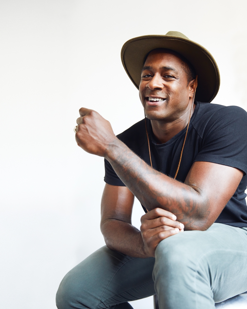 A young black man sits leaning-in in a relaxed and engaging pose with a big smile wearing a black t-shirt, jeans and wide-brimmed hat. 
