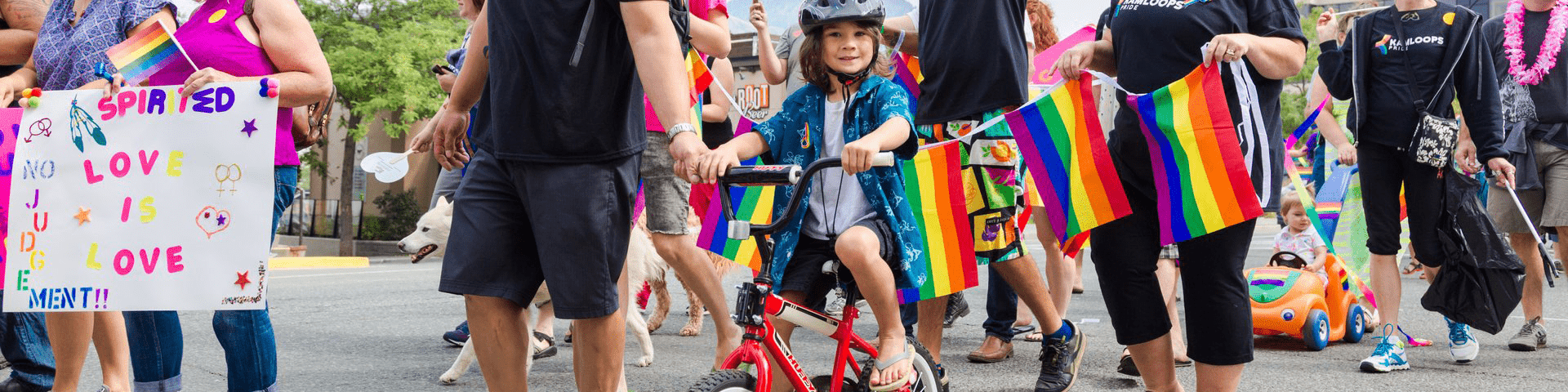 Young girl rides bike in 2017 Pride Parade in crowd of marchers carrying signs and rainbow flags