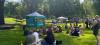 Residents sitting in the grass at Riverside Park watching a drag queen in a bright yellow dress give a presentation